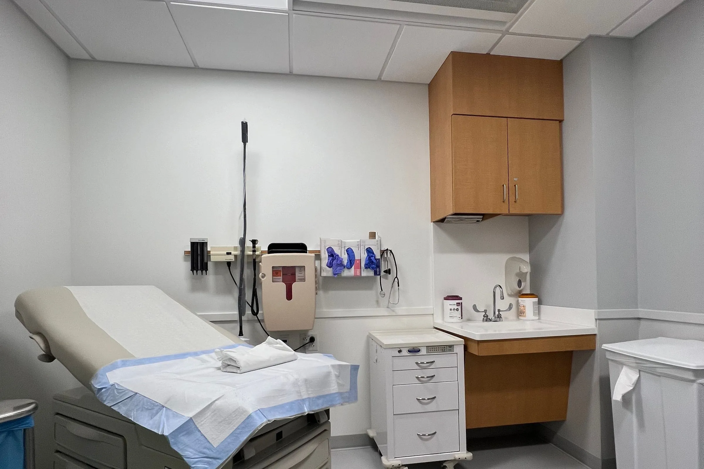 Exam room with a wood-grain upper cabinet, a sink counter with open knee space, and a drawer cart
